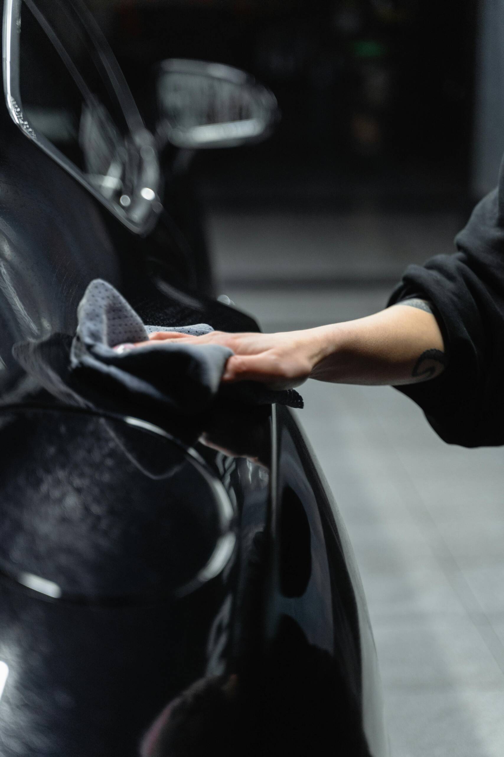 Close-up of a person's hand cleaning a shiny black car with a microfiber cloth in a garage.